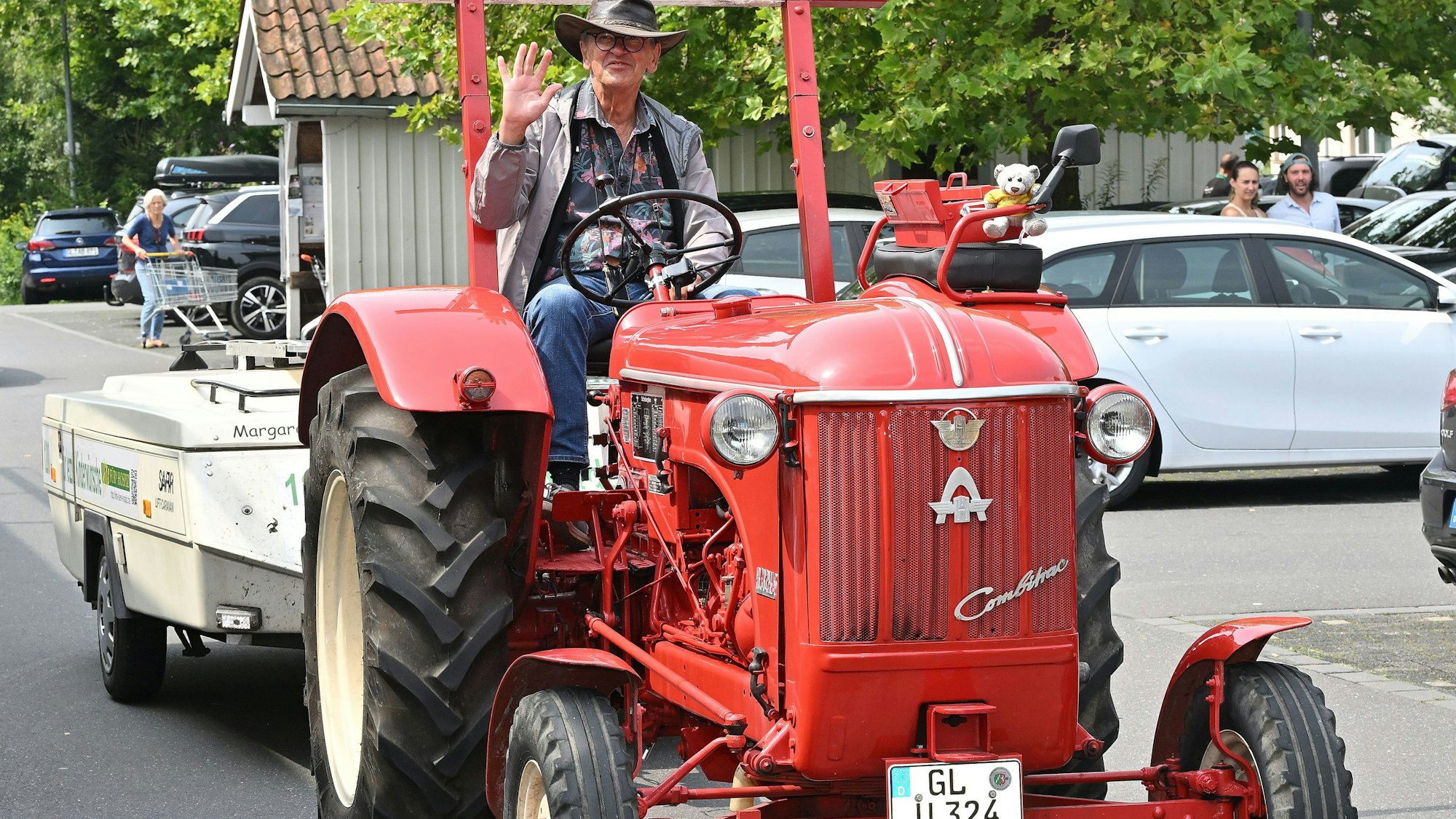 Ein Mann fährt auf einem Traktor mit Klappwohnanhänger und grüßt in die Kamera.