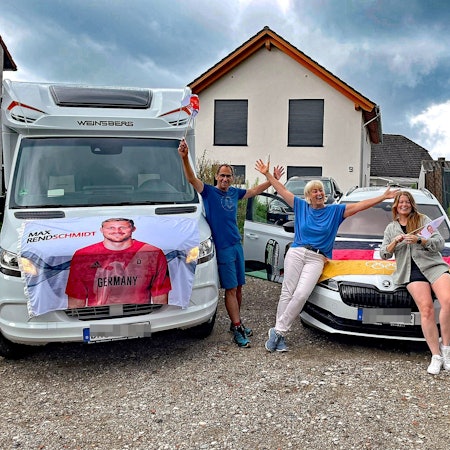 Ein Mann und zwei Frauen vor einem Wohnmobil und einem Pkw. Die Autos sind mit einem Transparent und einer Fahne geschmückt.