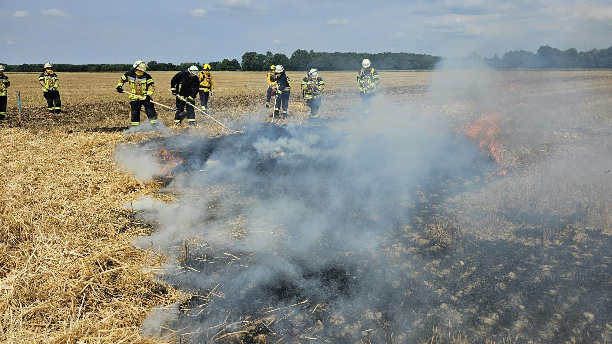 Dunkler Rauch liegt über einem brennenden Feld. Im Hintergrund sind Feuerwehrleute beschäftigt.