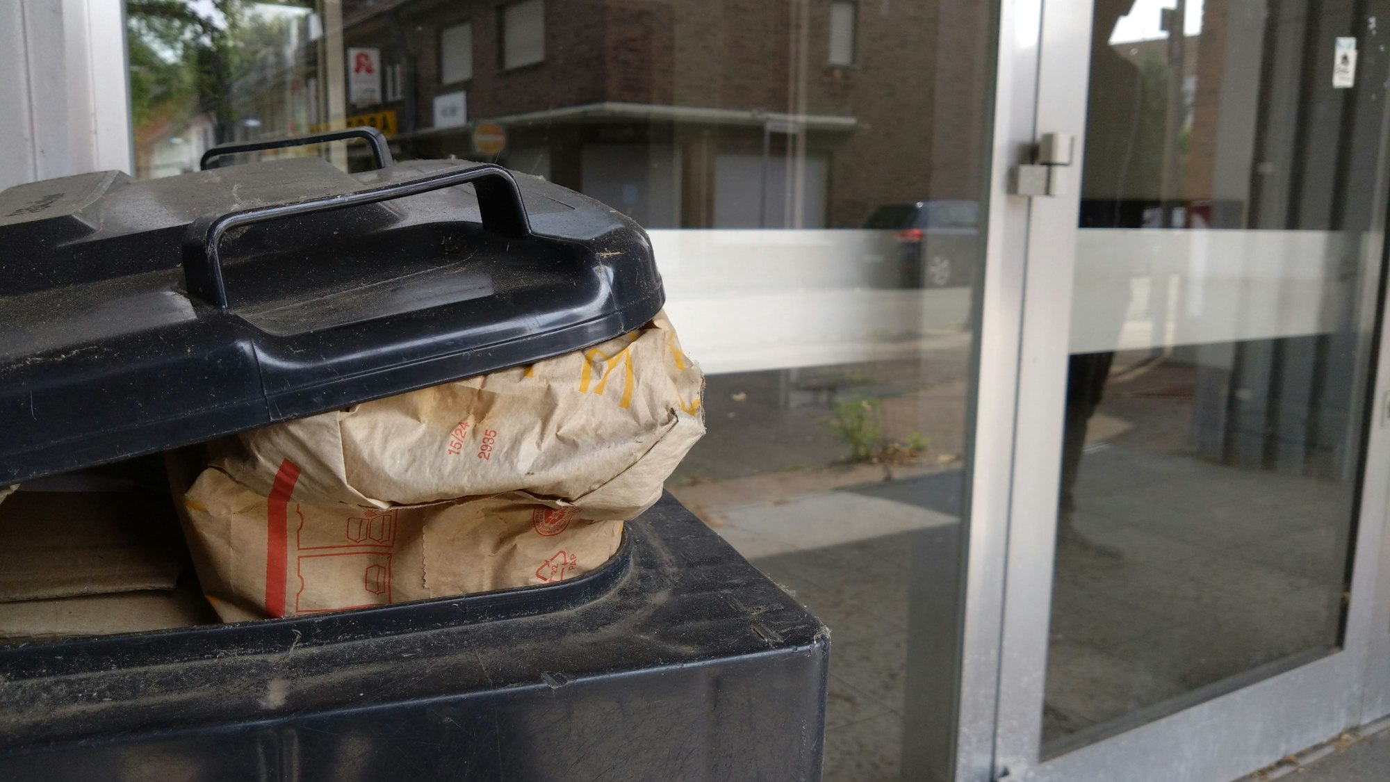 Die Türen des alten Rewe-Gebäudes, davor steht ein Mülleimer mit Fast-Food-Abfällen.