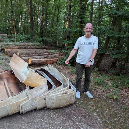 Ein Mann steht neben ineinander verschachtelten alten Kunststoffteilen vor einem Holzstapel auf einem Waldweg.