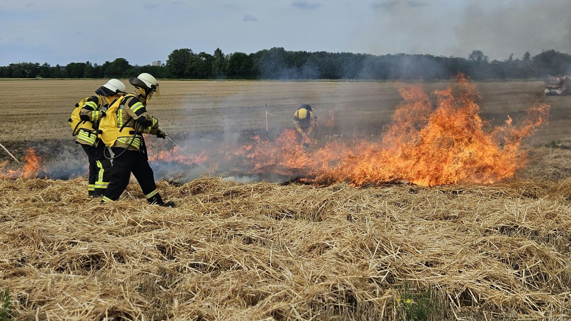 Feuerwehrleute stehen auf einem Strohfeld und löschen die Flammen eines Brandes.