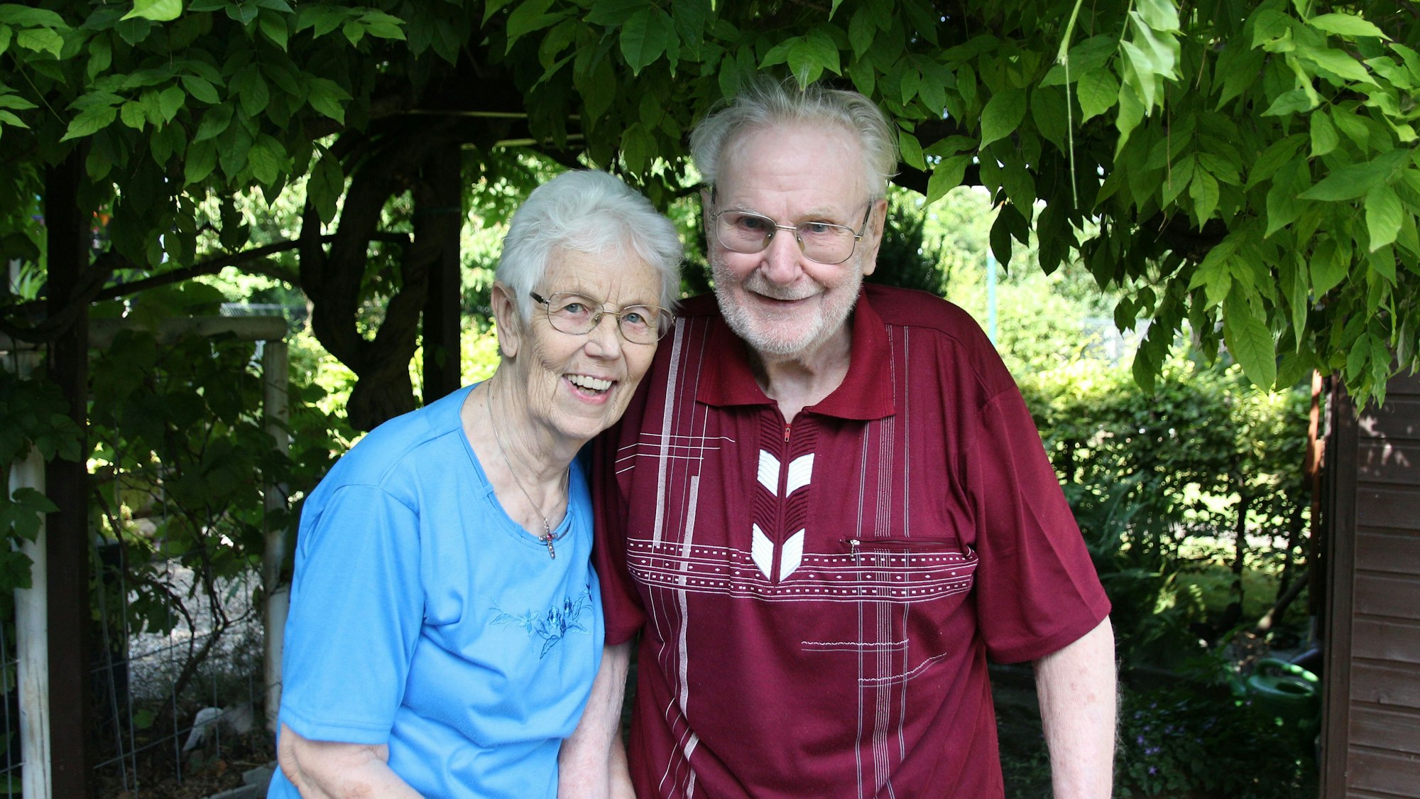 Das Pärchen lächelt in die Kamera. Fritz Jaeger trägt ein rotes Hemd mit weißen Mustern. Käthe Jaeger trägt eine hellblaue Bluse. Im Hintergrund einige Bäume.