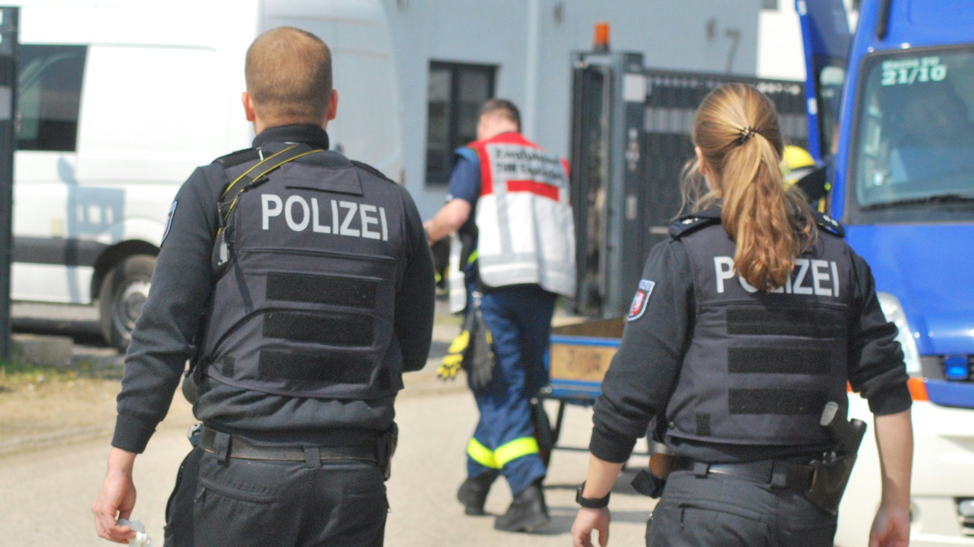 Zwei Polizisten gehen in Richtung einer Halle. Vor ihnen ist ein THW-Helfer zu erkennen.