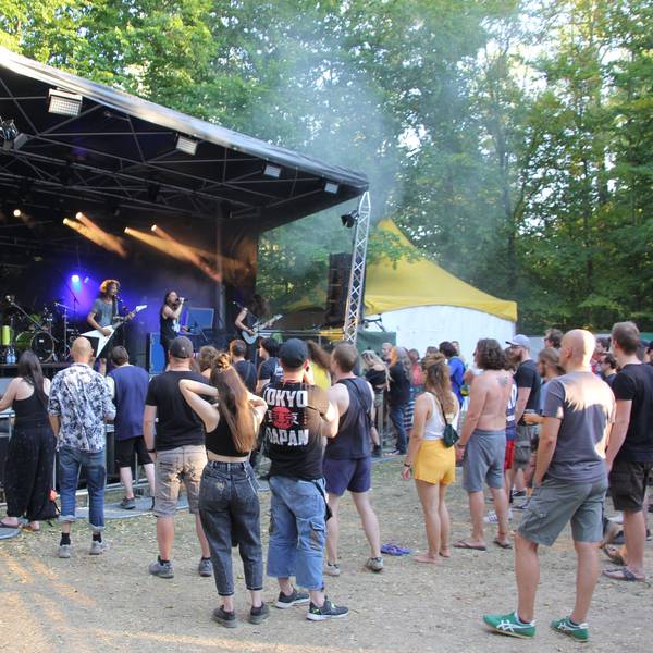Auf dem Bild ist die Bühne in Frechen zu sehen, eine Band tritt gerade auf. Vor der Bühne steht eine Menschenmenge. Im Hintergrund einige Bäume.