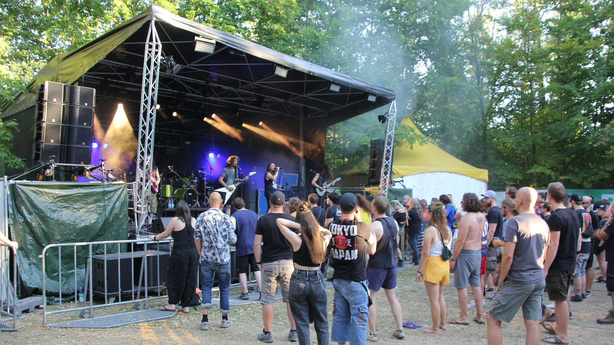 Auf dem Bild ist die Bühne in Frechen zu sehen, eine Band tritt gerade auf. Vor der Bühne steht eine Menschenmenge. Im Hintergrund einige Bäume.