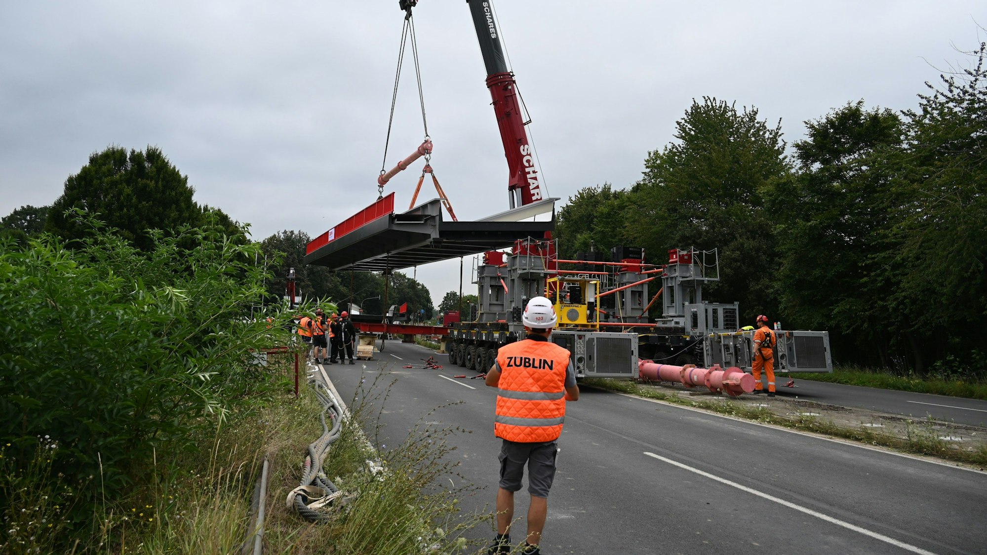 Ein Bauarbeiter in orangefarbener Warnweste beobachtet, wie das tonnenschwere Stahlbrückenteil mit dem Schwerlastkran verladen wird.