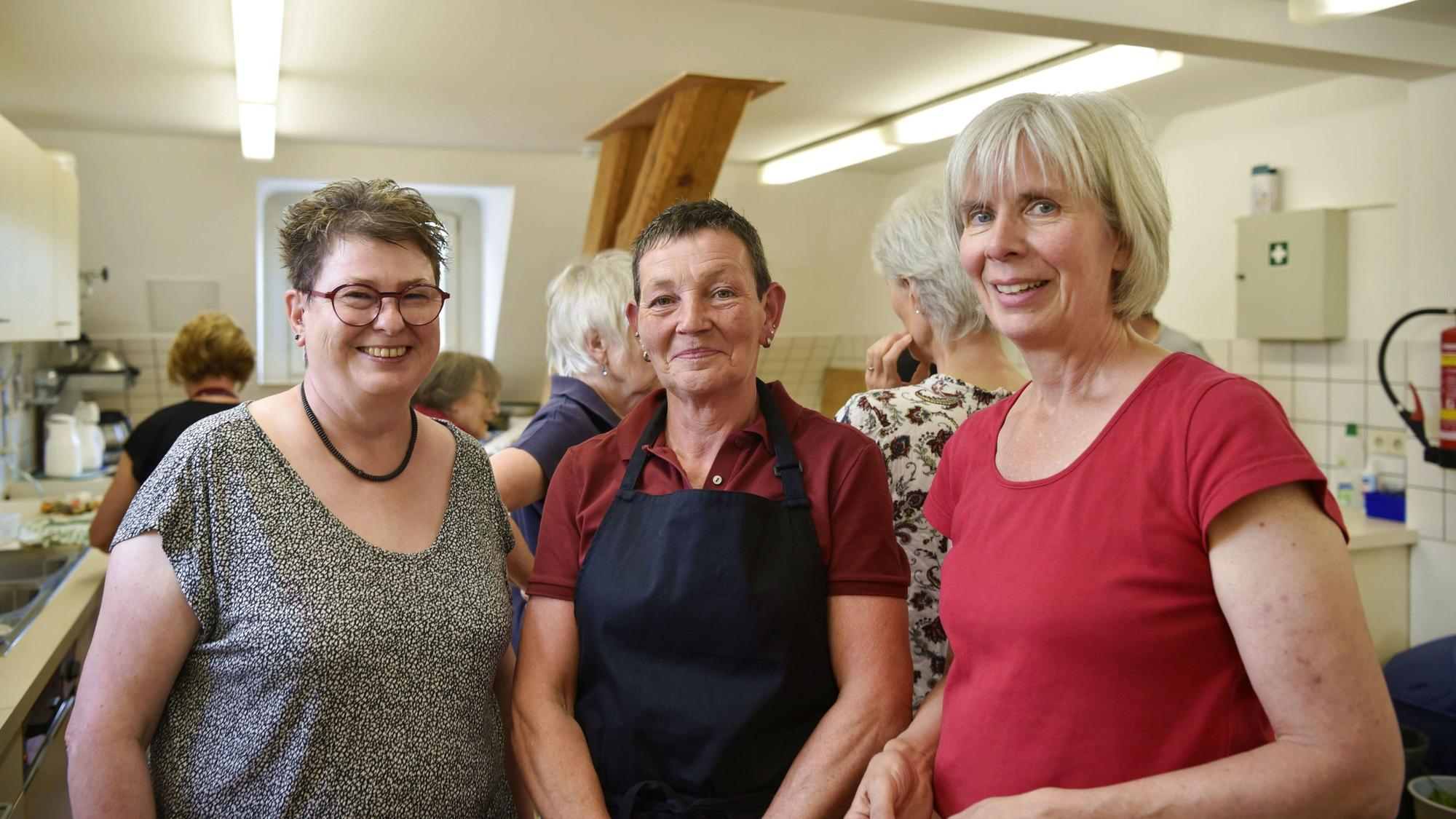 Helgard Hoeren (links) trägt ein schwarz-weißes T-Shirt, eine schwarze Kette, kurze Haare und eine rote runde Brille. Sie steht neben Petra Hartmann, die eine dunkelblaue Schürze über einem roten Poloshirt trägt. Daneben steht Ida Prinz-Hochgürtel, die ein rotes T-Shirt trägt. Im Hintergrund sind weitere Menschen und eine Küche zu erkennen.