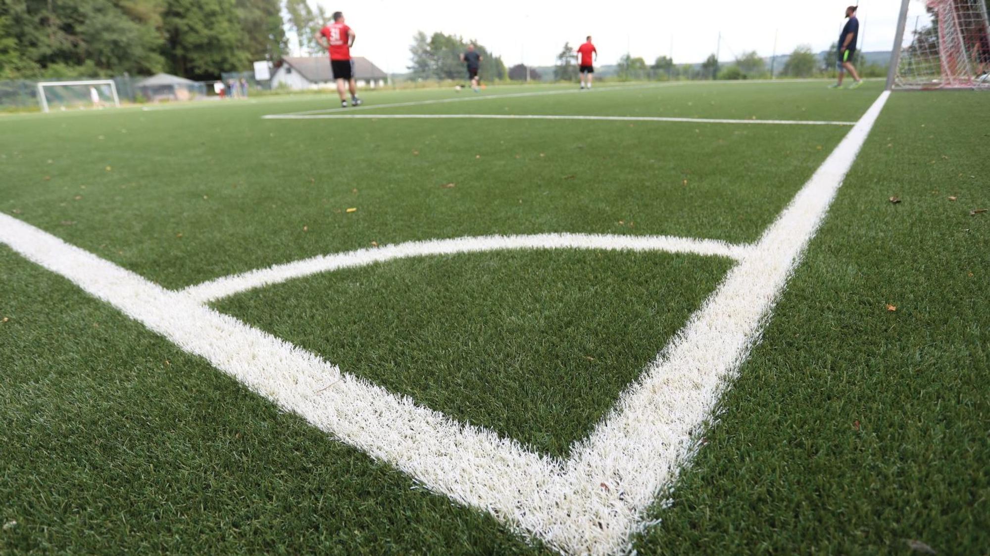 Ein Kleinspielfeld aus grünem Kunstrasen mit weißer Spielfeldmarkierung, im Hintergrund einige Fußballer.