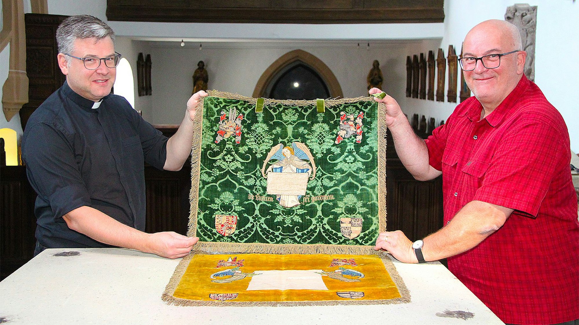 Zwei Männer, Pfarrer Andres Züll (links im schwarzen Hemd) und Markus Schmitz (rechts im roten Hemd) halten in der Pfarrkirche von Blankenheim zwei Tuchreliquien fest, die zum Kirchenschatz gehören.