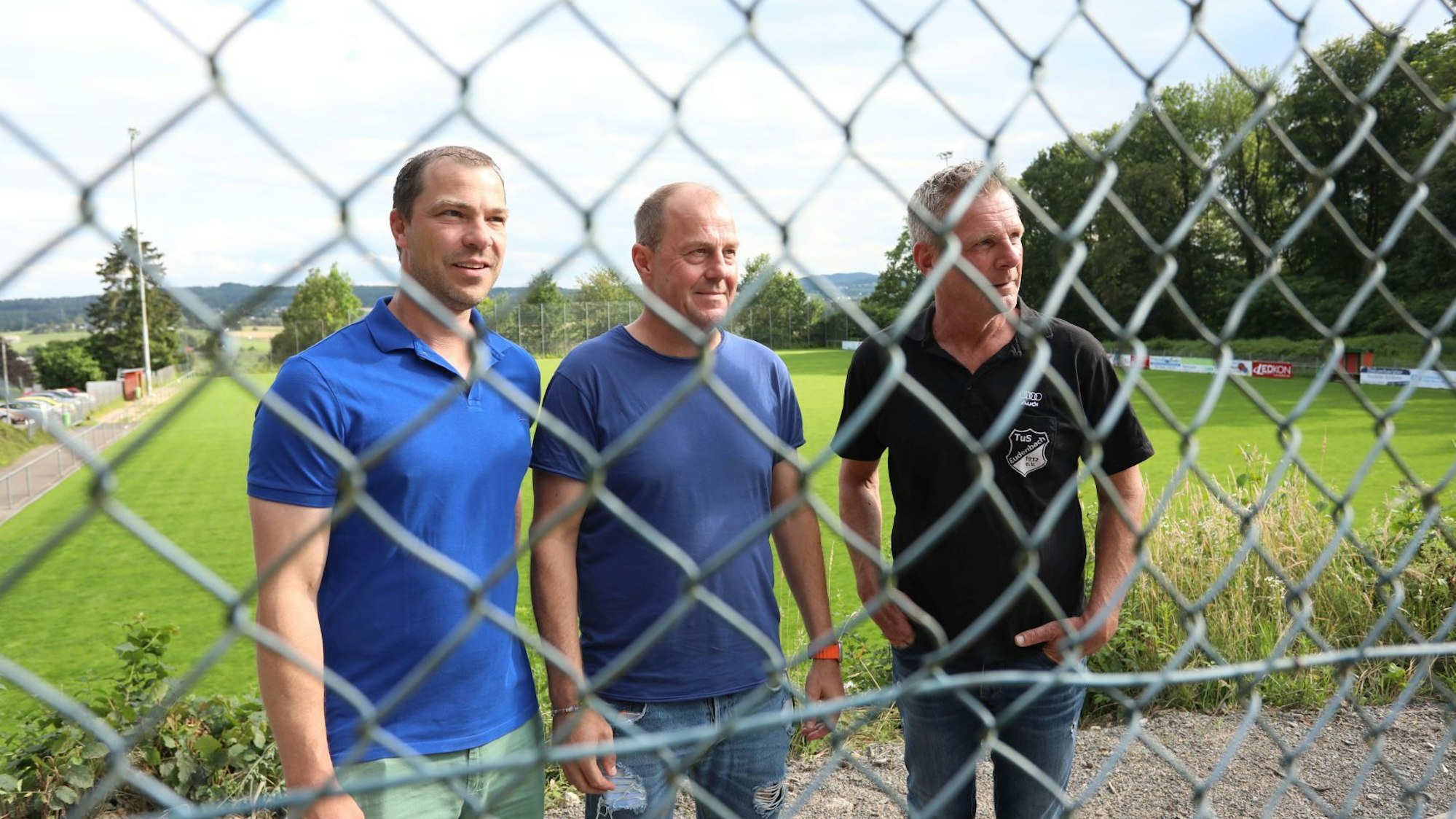 Drei Männer hinter einem Maschendrahtzaun, im Hintergrund der grüne Rasen eines Fußballplatzes.