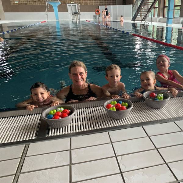 Die Schwimmlehrerin Carole Schifflers ist mit einigen Kindern im Wasser, am Beckenrand. Vor sich haben sie Schüsseln mit bunten Bällen. Im Hintergrund das Schwimmbad.