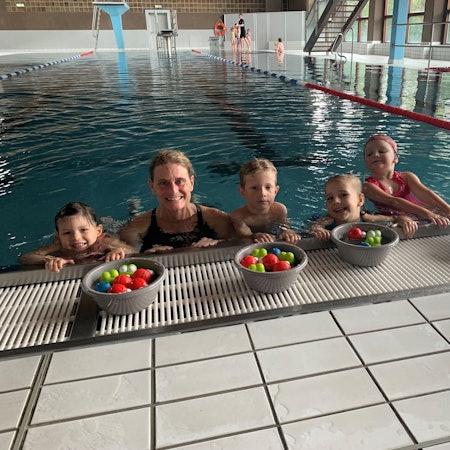 Die Schwimmlehrerin Carole Schifflers ist mit einigen Kindern im Wasser, am Beckenrand. Vor sich haben sie Schüsseln mit bunten Bällen. Im Hintergrund das Schwimmbad.
