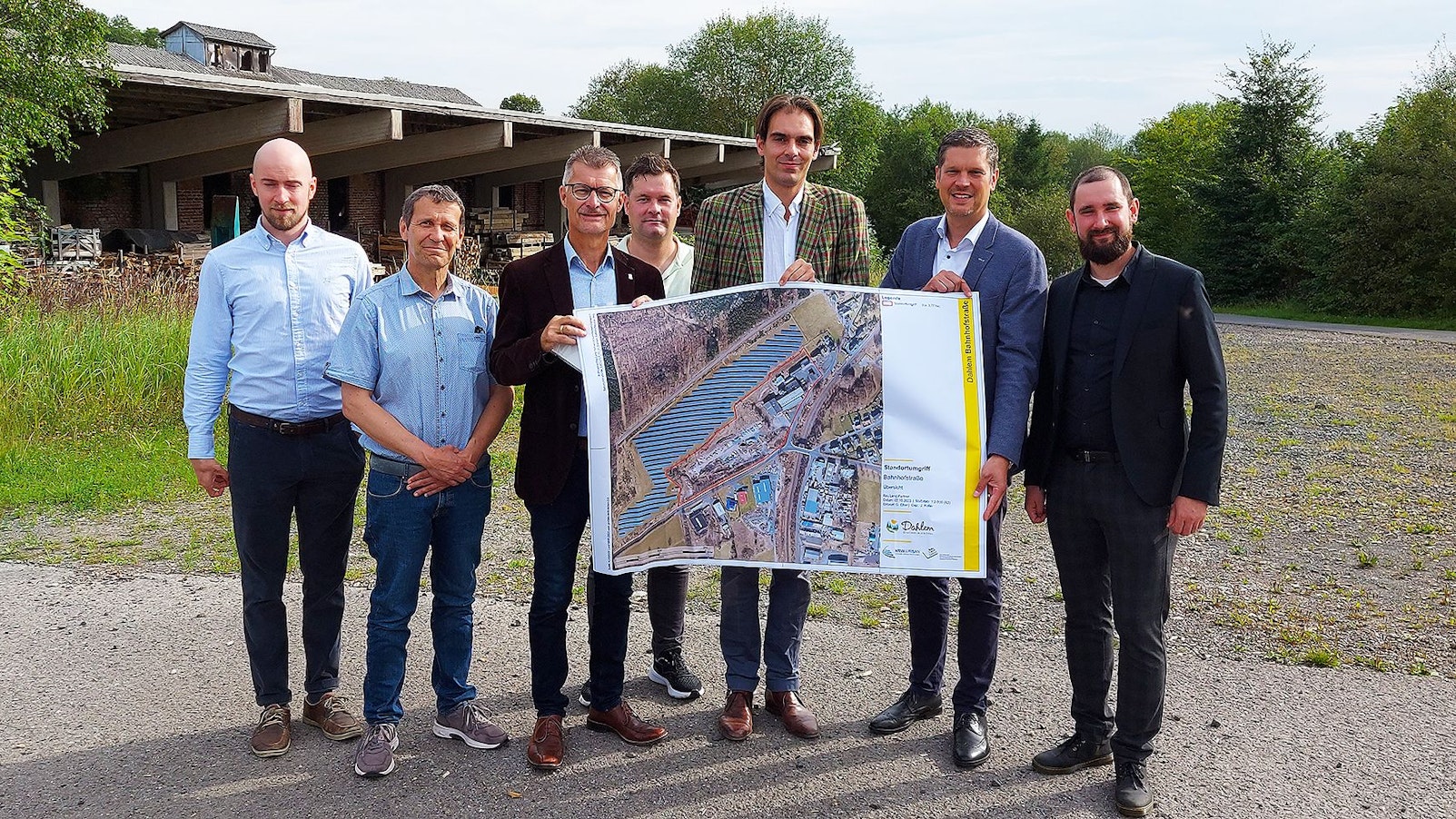 Eine Gruppe Männer steht auf dem Gelände des Sägewerks, einige halten einen Plan.