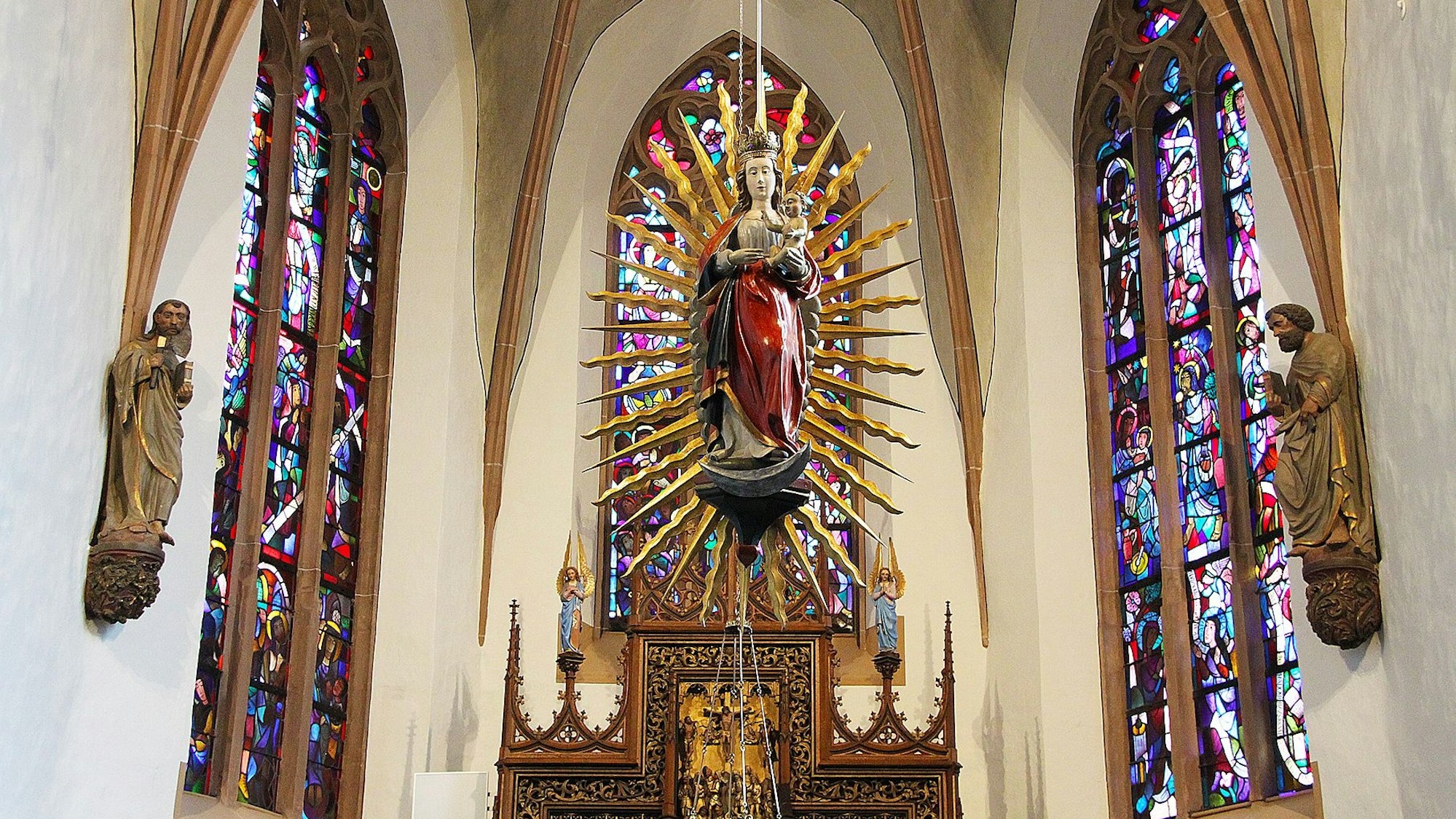 Im Altarraum der Blankenheimer Pfarrkirche ist eine Strahlenkranzmadonna zu sehen. Sie glänzt im Sonnenlicht, das durch die bunten Chorfenster fällt.