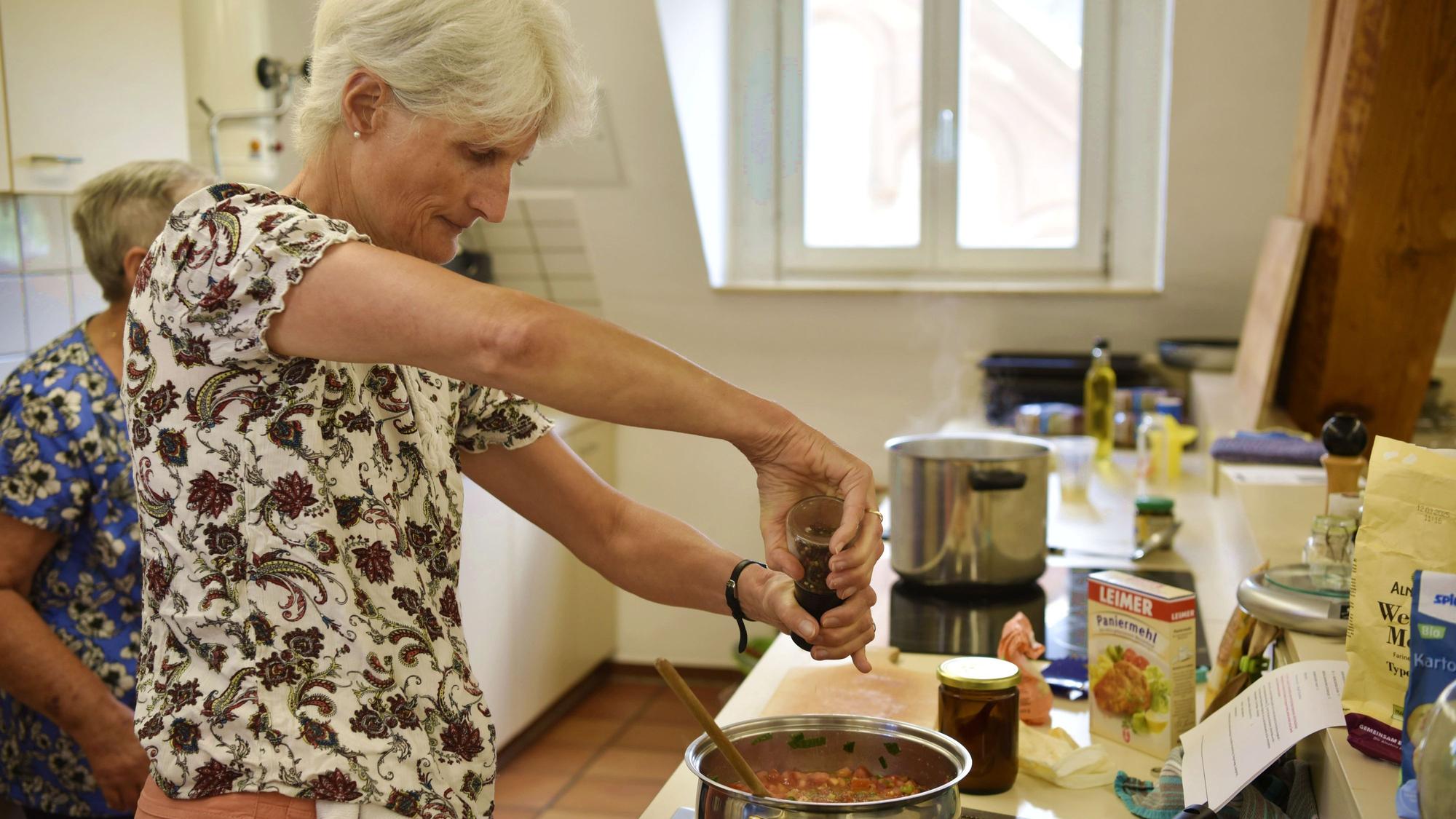 Elke Röhrig würzt eine in einem Topf vor sich hinkochende Tomatensoße mit Pfeffer. Sie trägt ein weißes T-Shirt mit roten Blumen.