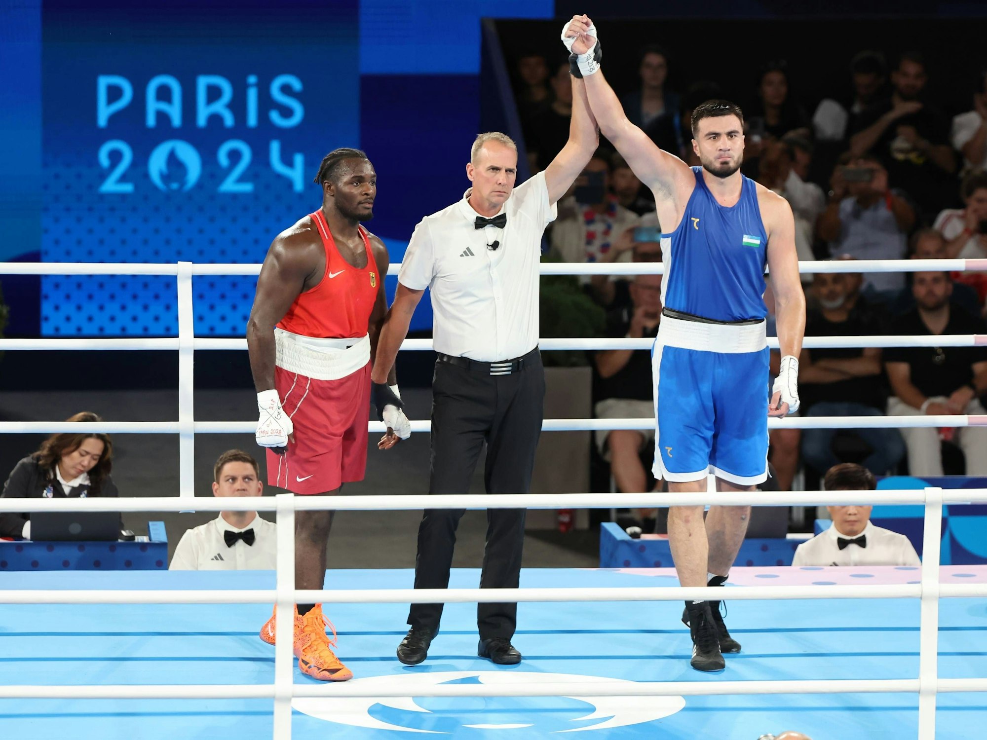 Das Bild zeigt Nelvie im Boxring mit seinem Gegner Bakhodir Jalolov aus Usbekistan und dem Ringrichter.