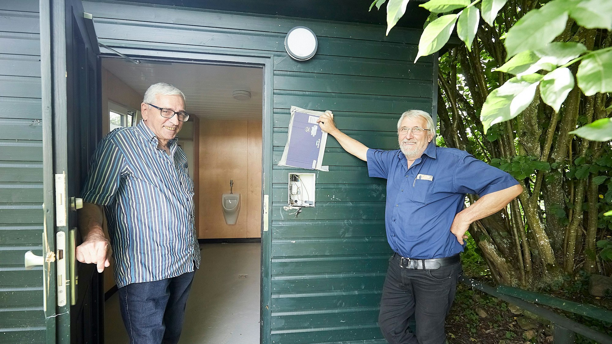 Heinz Schmitz und Gerhard Mayer stehen am Eingang des Toilettenhäuschens. Dort ist der Münzautomat aus der Wand gerissen.