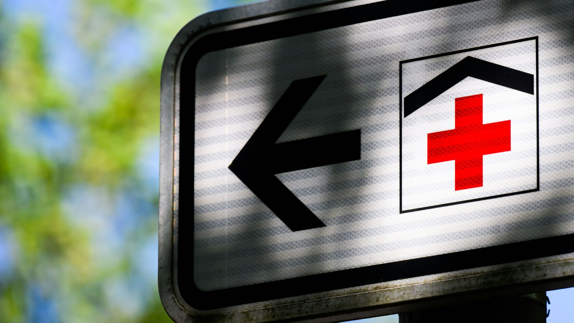 Ein Piktogramm mit einem roten Kreuz auf einem Straßenschild weist den Weg zu einem Krankenhaus.
