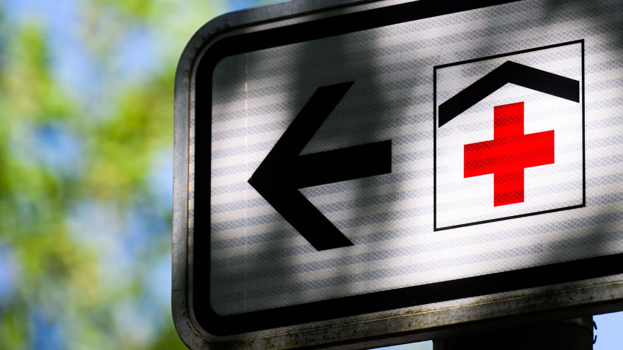 Ein Piktogramm mit einem roten Kreuz auf einem Straßenschild weist den Weg zu einem Krankenhaus.