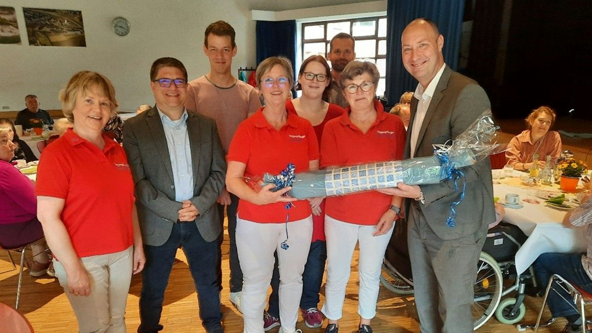 Das Bild zeigt einen Aufenthaltsraum des Haus Sonnenschein. Die Pflegerinnen sind in roten Hemden gekleidet und bekommen zur Feier ein Geschenk überreicht. (v.l.): Christina Fülop (Tagespflege), Bastian Bauer (stv. Gesamtleiter des CJD-Verbunds NRW Süd), Carsten Zanchettin (Tagespflege), Ute Repp (Leiterin Tagespflege), Andrea Winkel (Tagespflege), Sebastian Reuß (Fachbereichsleiter CJD Frechen), Margit Albrecht (Tagespflege) und Boris Lietz (Gesamtleiter des CJD Verbund NRW Süd).