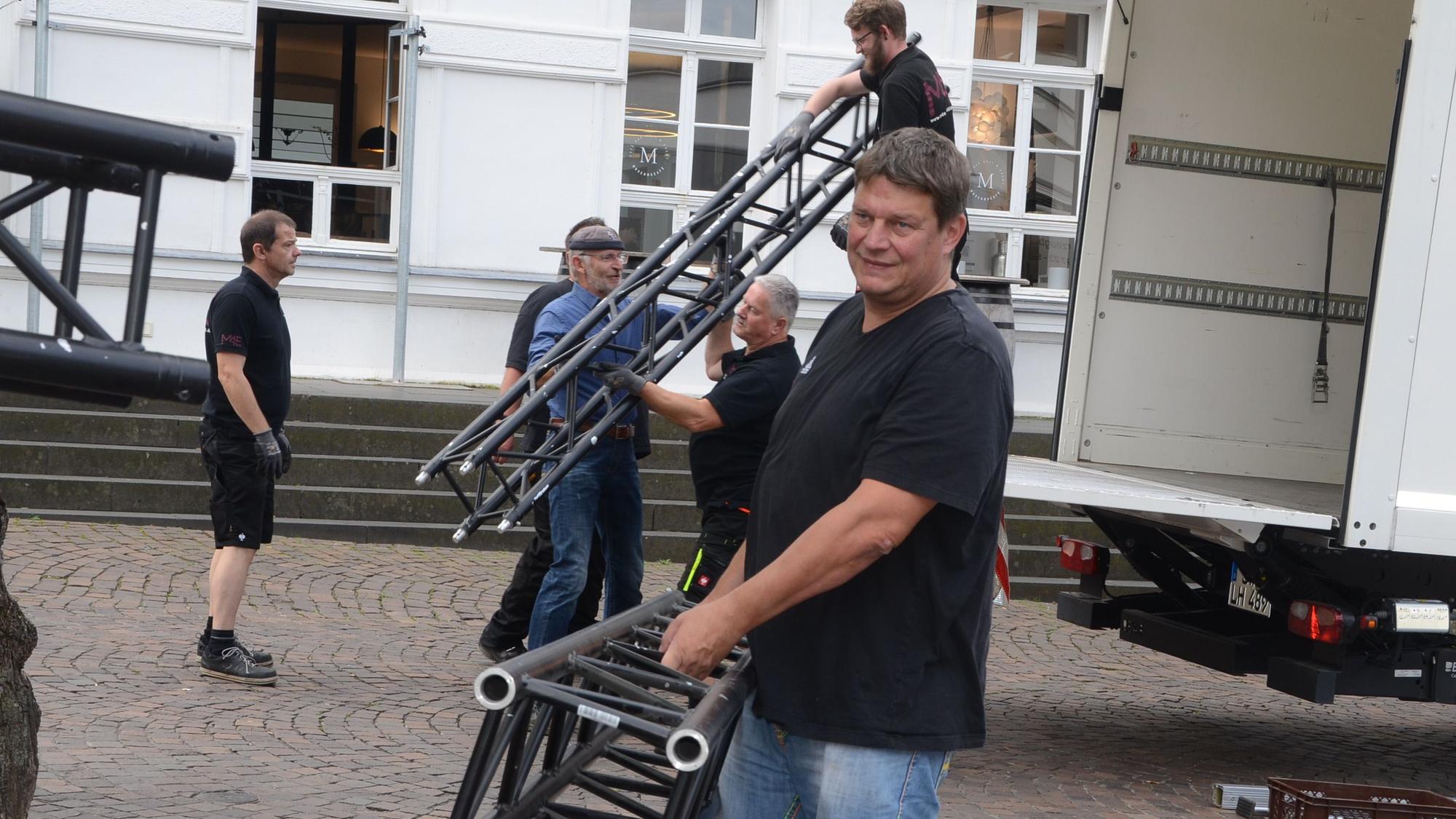 Ein Mann trägt einen Pfosten für eine Bühne.Aus einem Lastwagen werden weitere Bühnenteile herausgehoben.