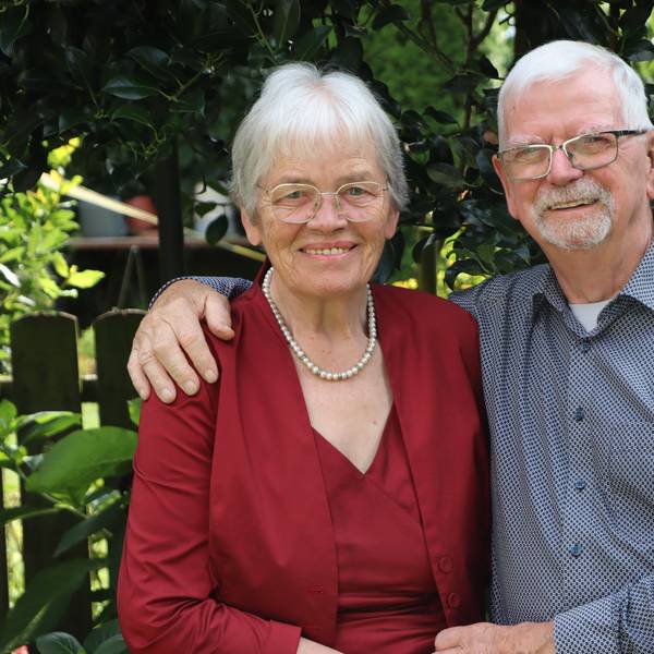 Ursula Abeln trägt eine Brille, eine rote Bluse und eine Perlenkette. Gerhard Abeln hat seinen Arm um seine Frau gelegt, er trägt eine Brille und ein blaues, gepunktetes Hemd. Im Hintergrund ein Garten.