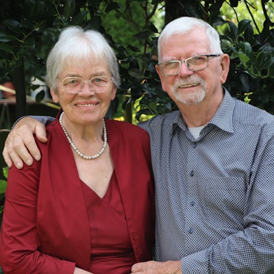 Ursula Abeln trägt eine Brille, eine rote Bluse und eine Perlenkette. Gerhard Abeln hat seinen Arm um seine Frau gelegt, er trägt eine Brille und ein blaues, gepunktetes Hemd. Im Hintergrund ein Garten.