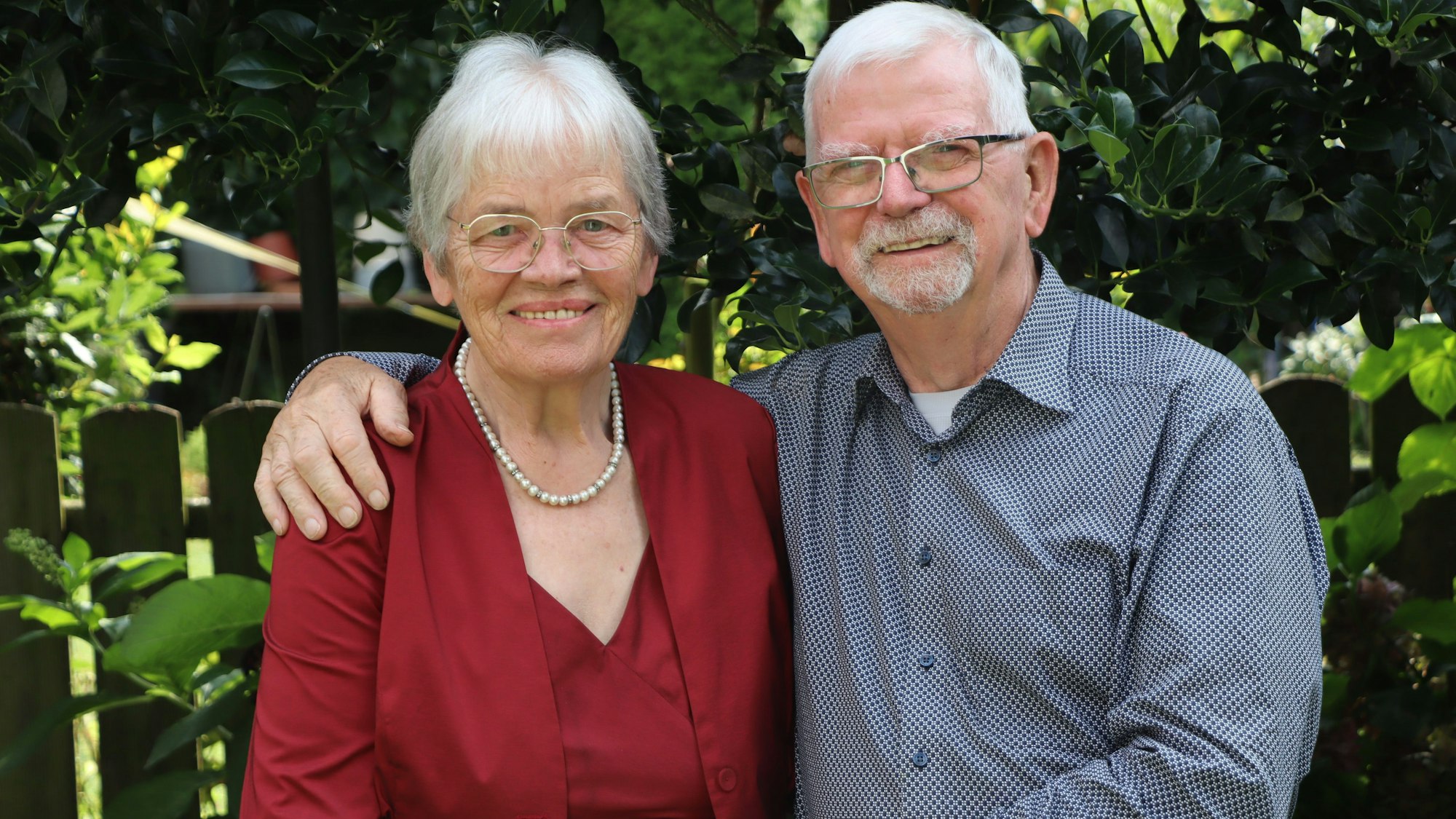 Ursula Abeln trägt eine Brille, eine rote Bluse und eine Perlenkette. Gerhard Abeln hat seinen Arm um seine Frau gelegt, er trägt eine Brille und ein blaues, gepunktetes Hemd. Im Hintergrund ein Garten.