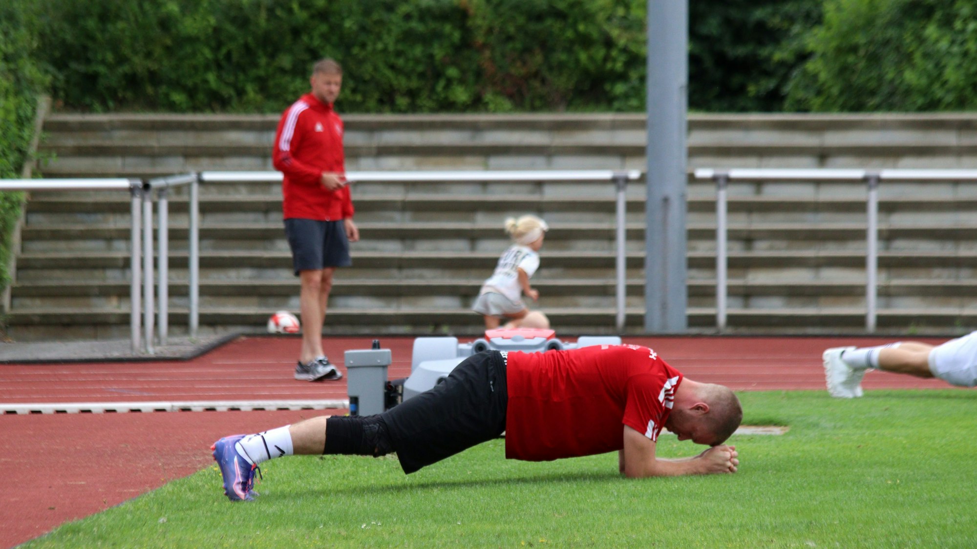 Ein Fußballtorwart plankt am Spielfeldrand. Im Hintergrund fährt ein Mädchen auf einem Roller über eine Tartanbahn.