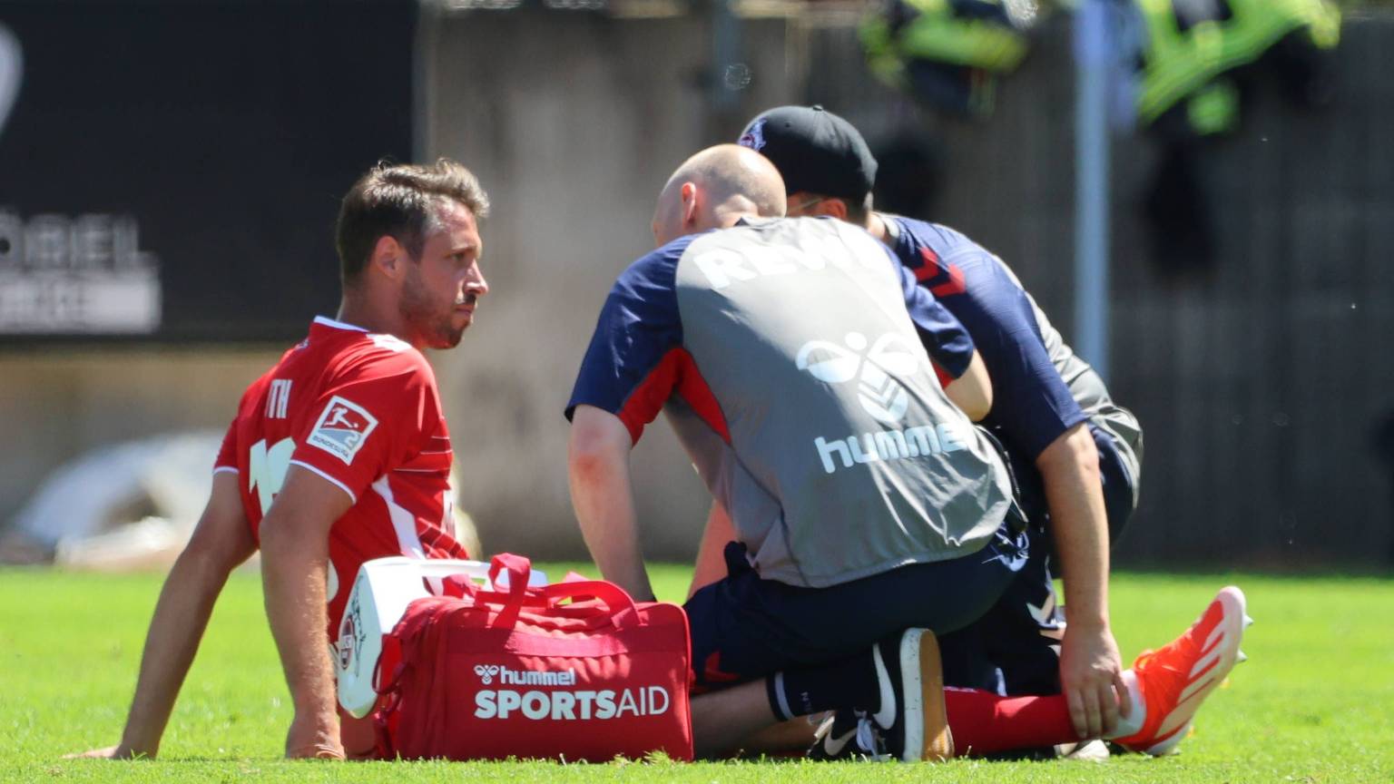 Mark Uth sitzt verletzt auf dem Rasen und bespricht sich mit den Physiotherapeuten des 1. FC Köln.