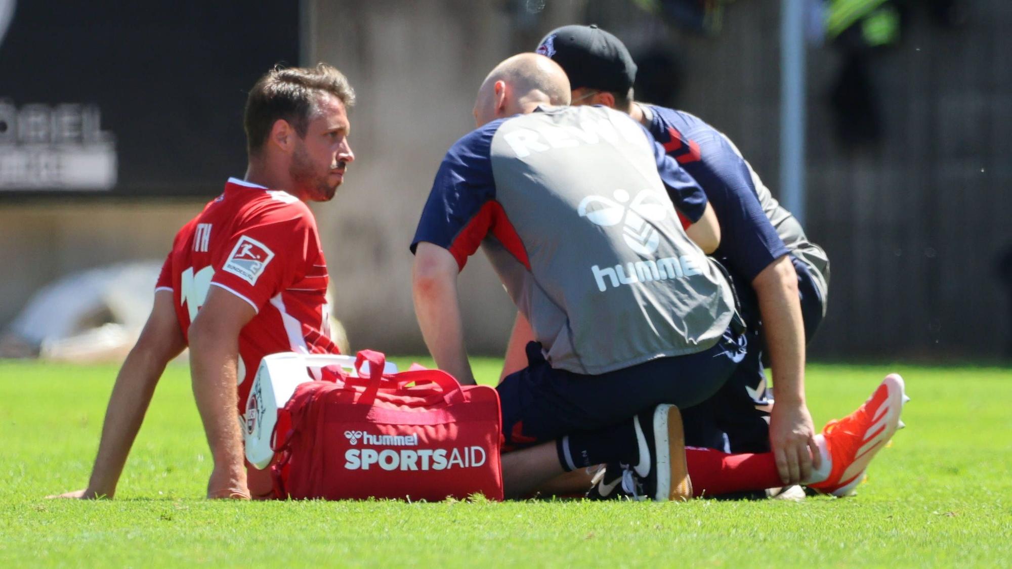 Mark Uth sitzt verletzt auf dem Rasen und bespricht sich mit den Physiotherapeuten des 1. FC Köln.