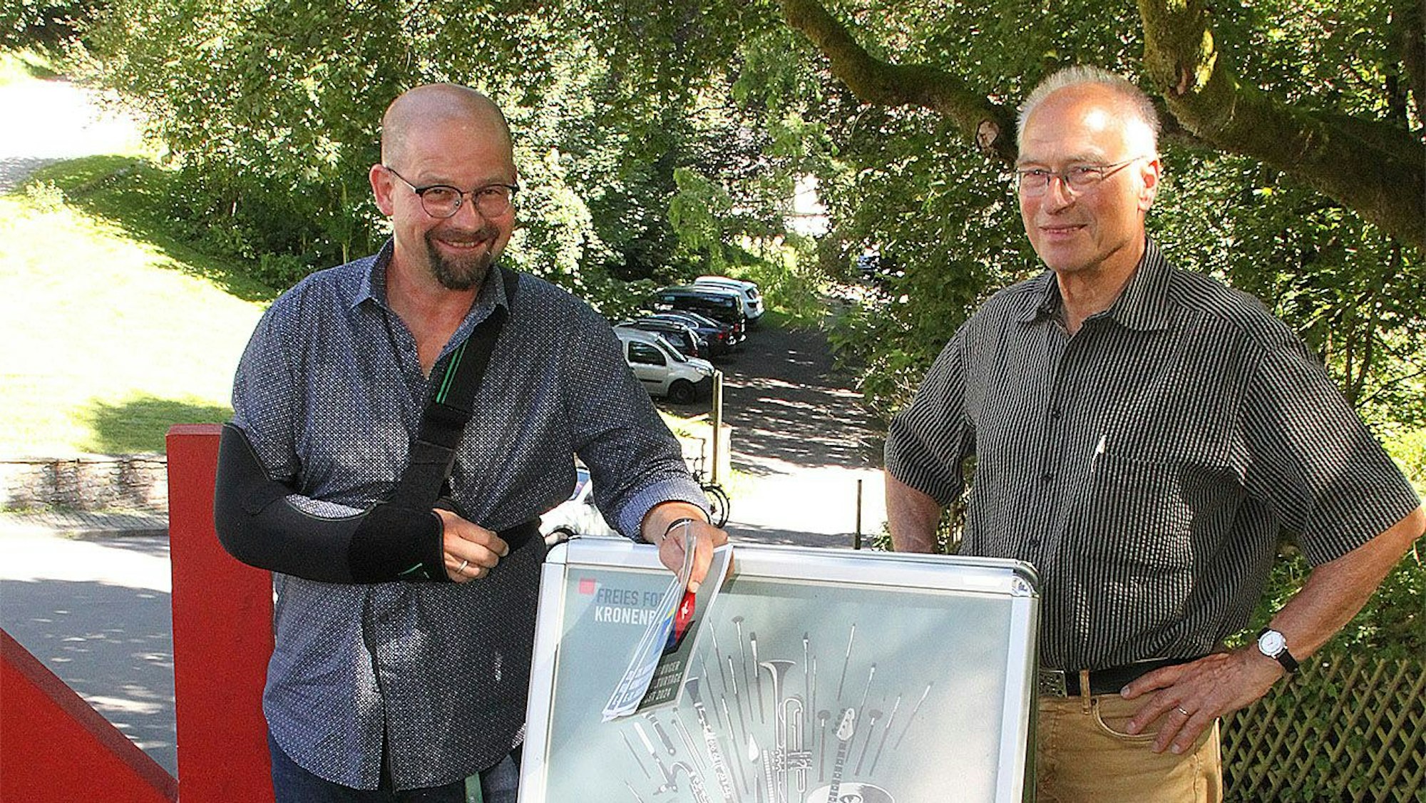 Zwei Männer, Martin Schöddert (links mit einer schwarzen Schulterschlinge) und Pit Igelmund, stehen an einem Plakataufsteller der Kronenburger Kunst- und Kulturtage.