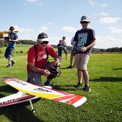 Ein Mann hockt neben einem Modellflugzeug. Er hält eine Fernbedienung in der Hand. Neben ihm steht ein Junge.