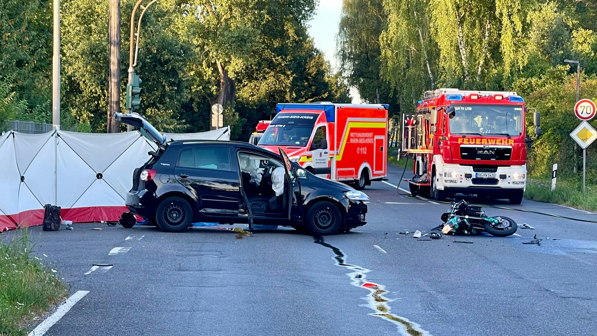 Ein Auto steht auf einer Straße vor mehreren Einsatzfahrzeugen der Feuerwehr, ein zerstörtes Motorrad liegt auf der Straße. Ein Sichtschutz ist hinter dem Pkw aufgebaut.