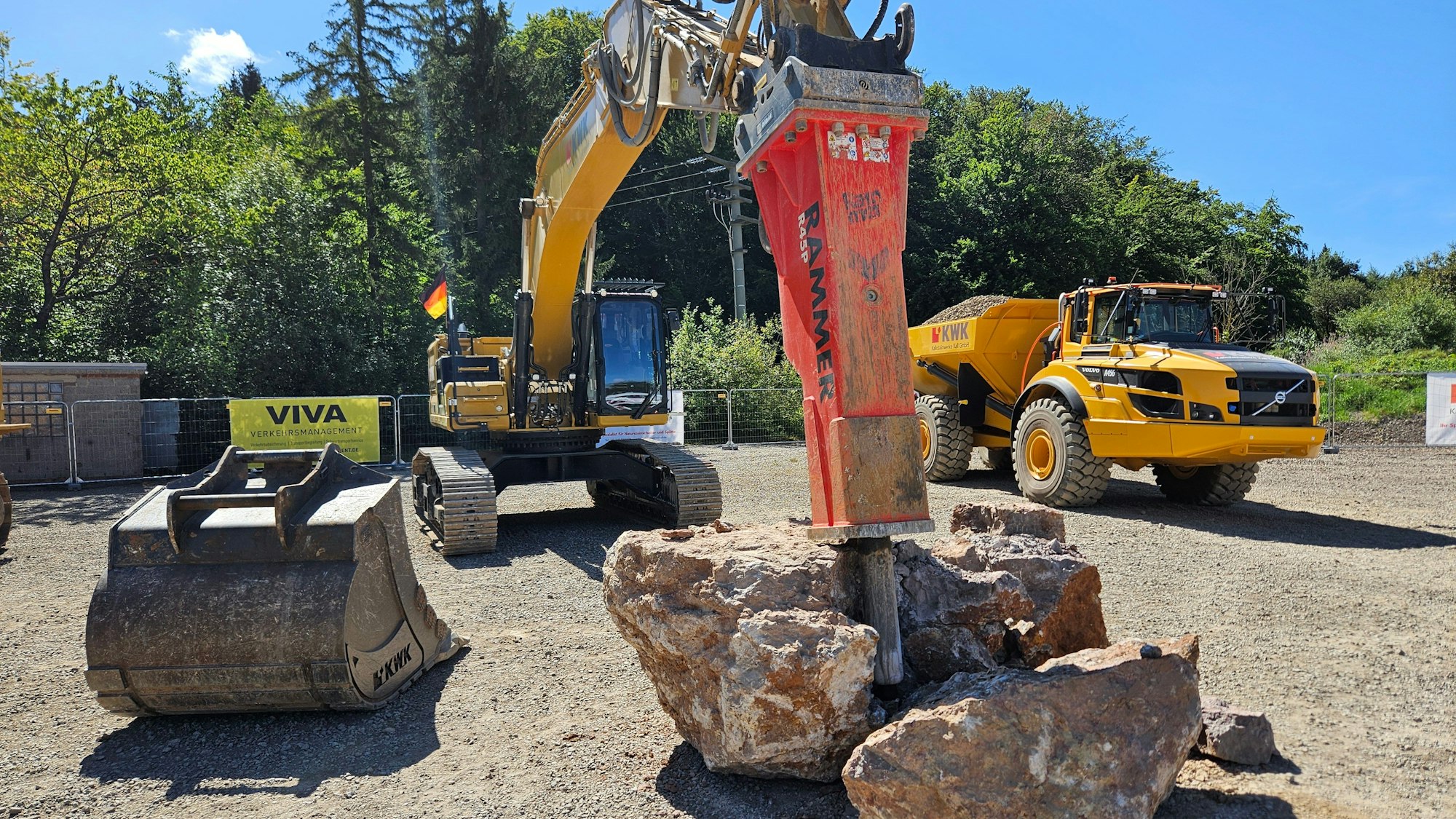 Ein Bagger mit einem Hydraulikhammer ist zu sehen. Daneben steht ein beladener Muldenkipper.