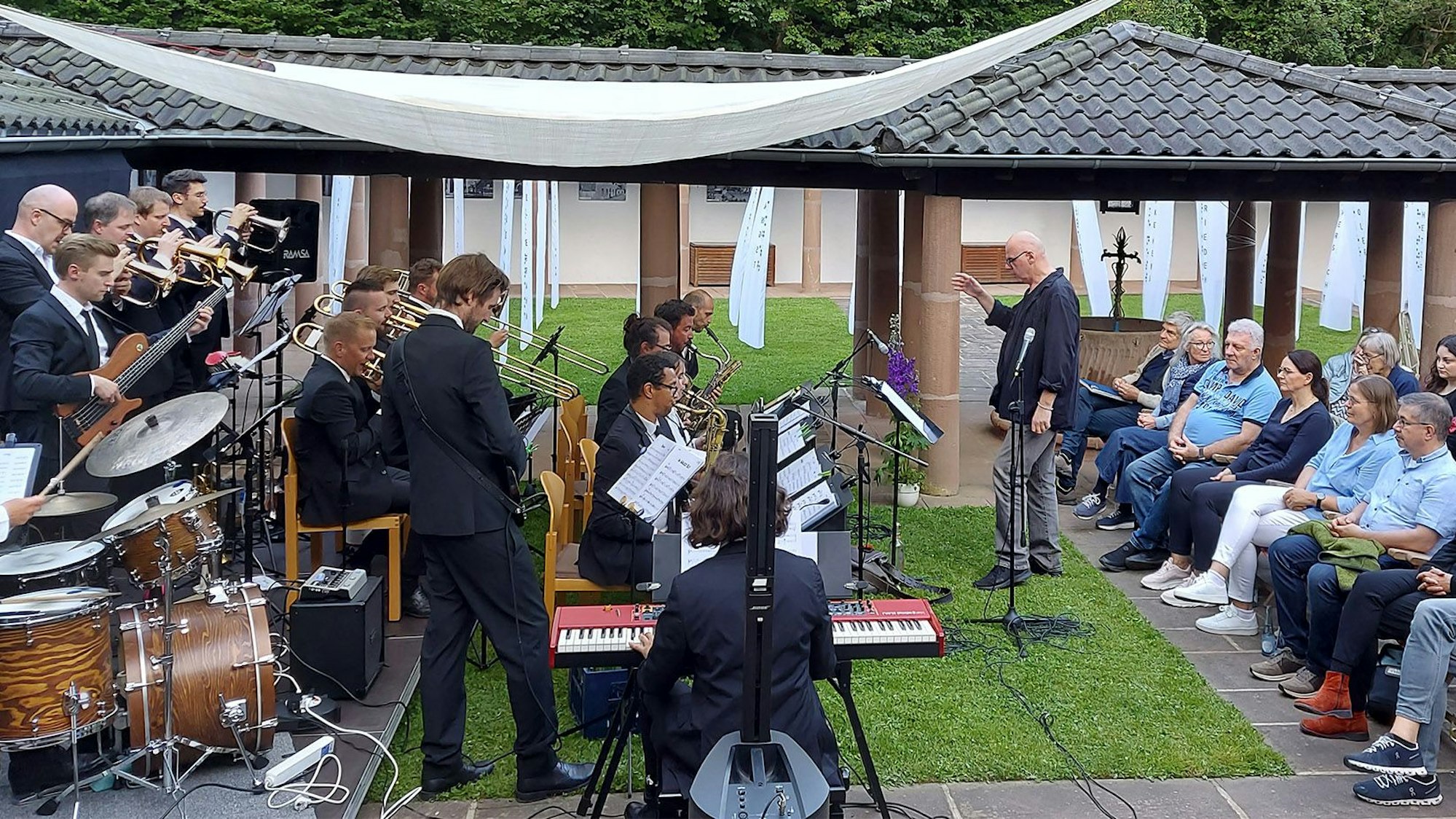 Frank Reinshagen steht in der Mitte und dirigiert ein Oechester, das links zu sehen. Rechts ist ein Teil des Publikums in einem Innenhof in Dahlem-Kronenburg zu sehen.