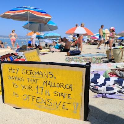 El Arenal: ‚Hallo Deutsche, zu sagen, dass Mallorca euer 17. Bundesland ist, ist offensiv-aggressiv‘ steht auf einem Schild, das an einem Strand auf Mallorca aus Protest gegen den Massentourismus auf der Balearen-Insel gezeigt wird.