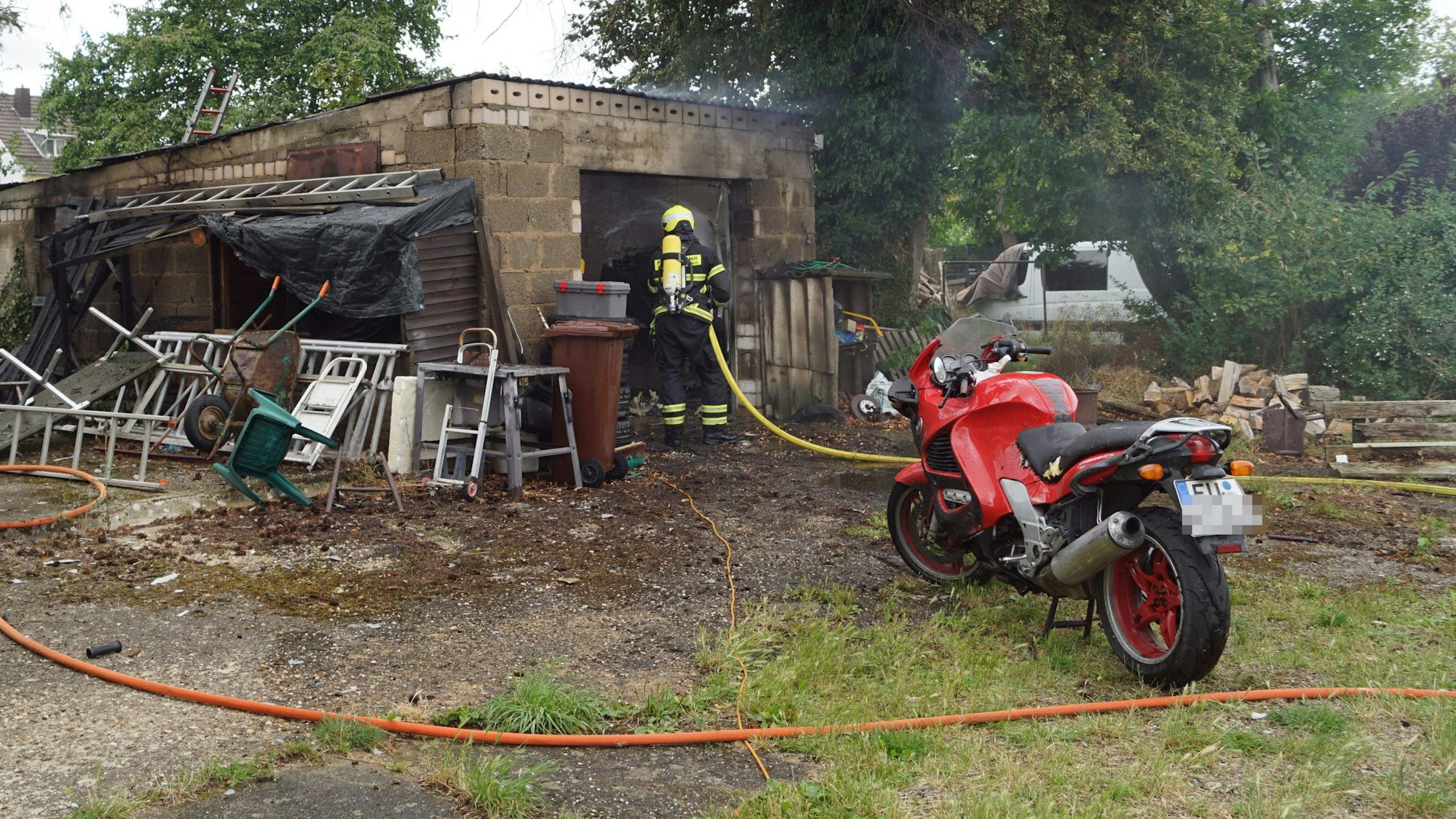 Eine Einsatzkraft der Feuerwehr ist mit einem Atemschutzgerät ausgerüstet und löscht einen Brand in einer Garage. Vor der Garage steht ein rotes Motorrad.