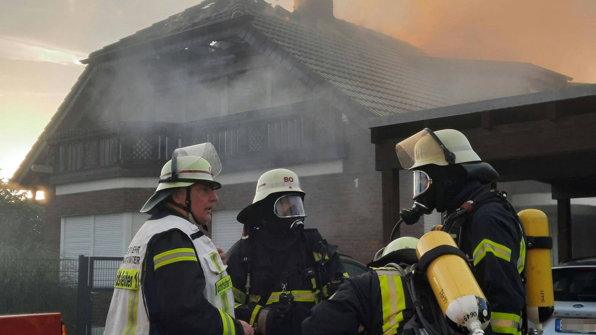 Feuerwehrleute mit Atemschutzgeräten stehen vor einem brennenden Haus.