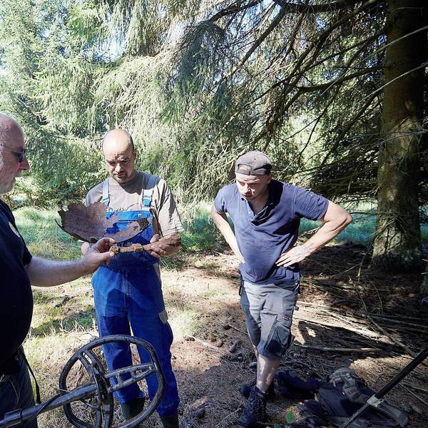 Drei Männer, Gerald Scholz, Mark Michels und Ramon Bügler, stehen in einem Wald. Einer hat ein Metallsuchgerät in der Hand. Sie schauen sich verrostete Metallteile an, bei denen es sich um Teile einer V1-Bombe aus dem Zweiten Weltkrieg handelt.