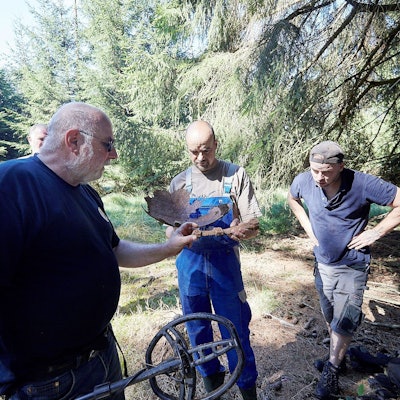 Drei Männer, Gerald Scholz, Mark Michels und Ramon Bügler, stehen in einem Wald. Einer hat ein Metallsuchgerät in der Hand. Sie schauen sich verrostete Metallteile an, bei denen es sich um Teile einer V1-Bombe aus dem Zweiten Weltkrieg handelt.