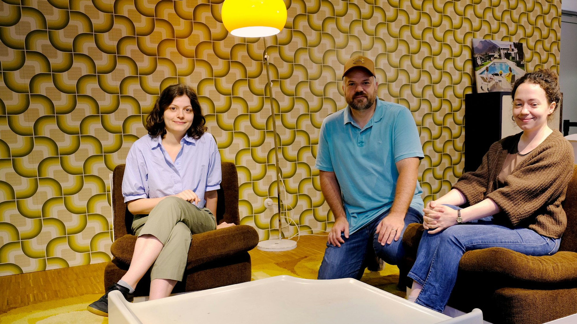 Clara Ruf (l.) und Jennifer Gnädinger (hier im Bild mit Veranstaltungs-Manager Daniel Manner) sitzen inmitten eines Wohnzimmers im 70er-Jahre-Stil. Teppich und Tapete sind bunt gemustert.