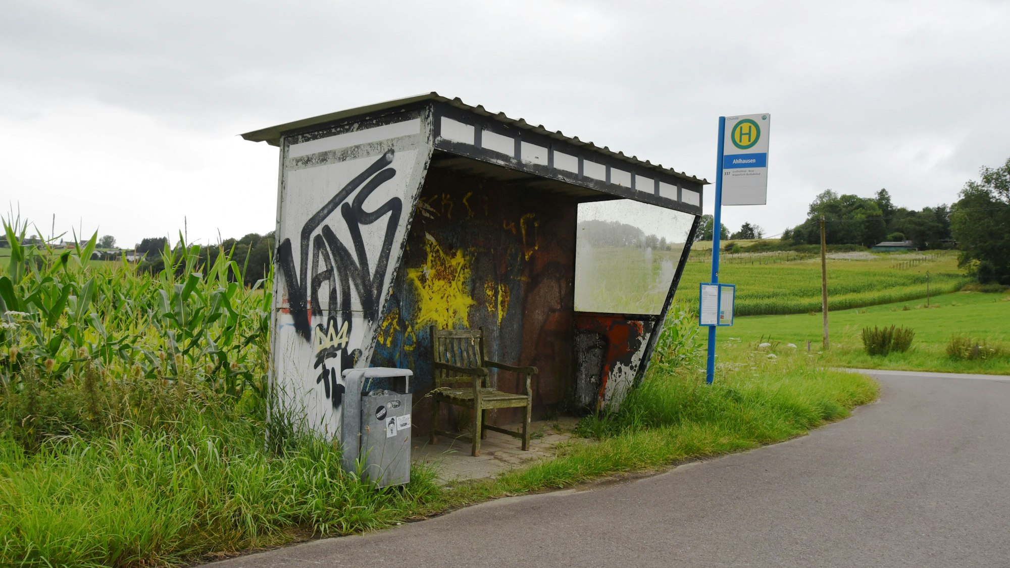 Eine mit Graffiti versehene Bushaltestelle steht mitten im Nirgendwo, drumherum nur Felder und weite Landschaften. Ein Haltestellenschild steht aus Sicht des Betrachtenden rechts von der Haltestelle. Links von der Haltestelle steht ein Mülleimer.