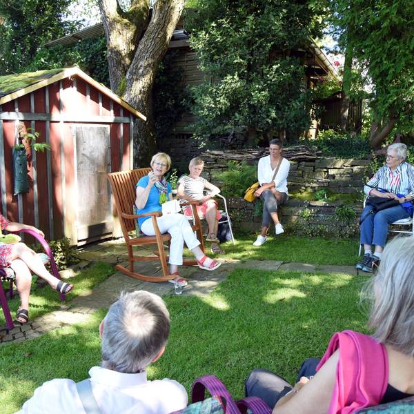 Das Foto zeigt eine Gruppe von Menschen, die in einem Gartensitzen und der Vorleserin Gisela Osenberg lauschen.