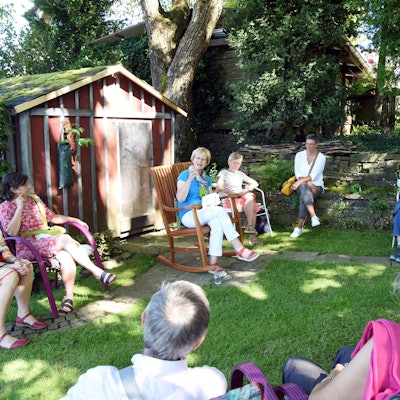 Das Foto zeigt eine Gruppe von Menschen, die in einem Gartensitzen und der Vorleserin Gisela Osenberg lauschen.