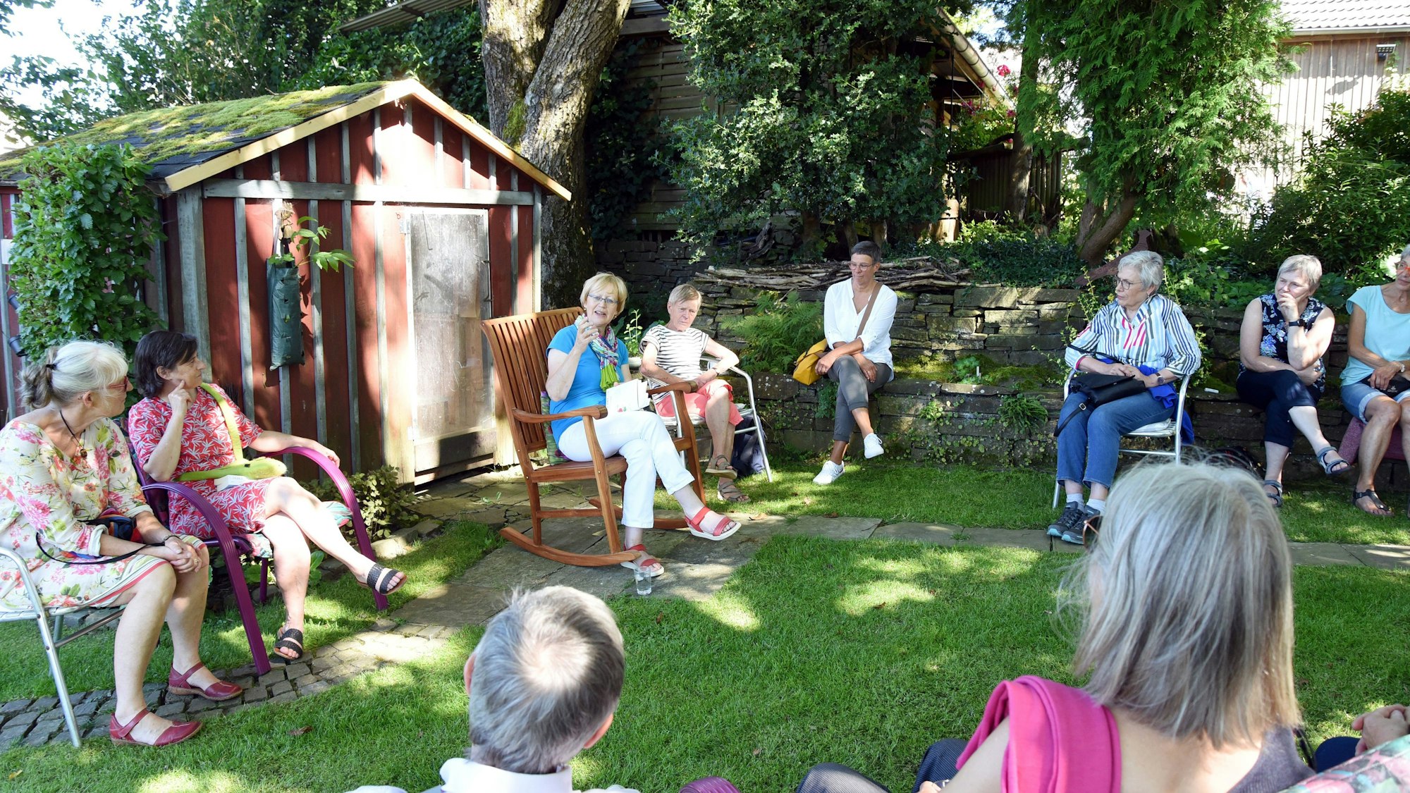 Das Foto zeigt eine Gruppe von Menschen, die in einem Gartensitzen und der Vorleserin Gisela Osenberg lauschen.