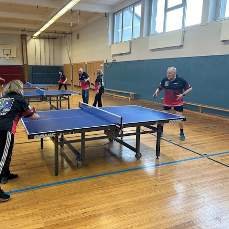 Mehrere Leute spielen in einer Sporthalle an mehreren Platten Tischtennis.