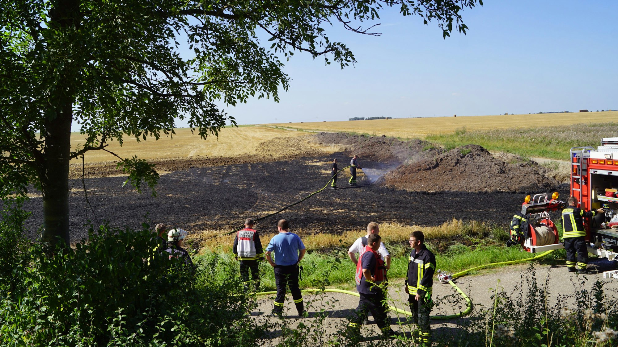 Auf einem Feld qualmen Strohballen. Rechts im Bild ein Feuerwehrauto, Feuerwehrleute löschen mit einem Schlauch.