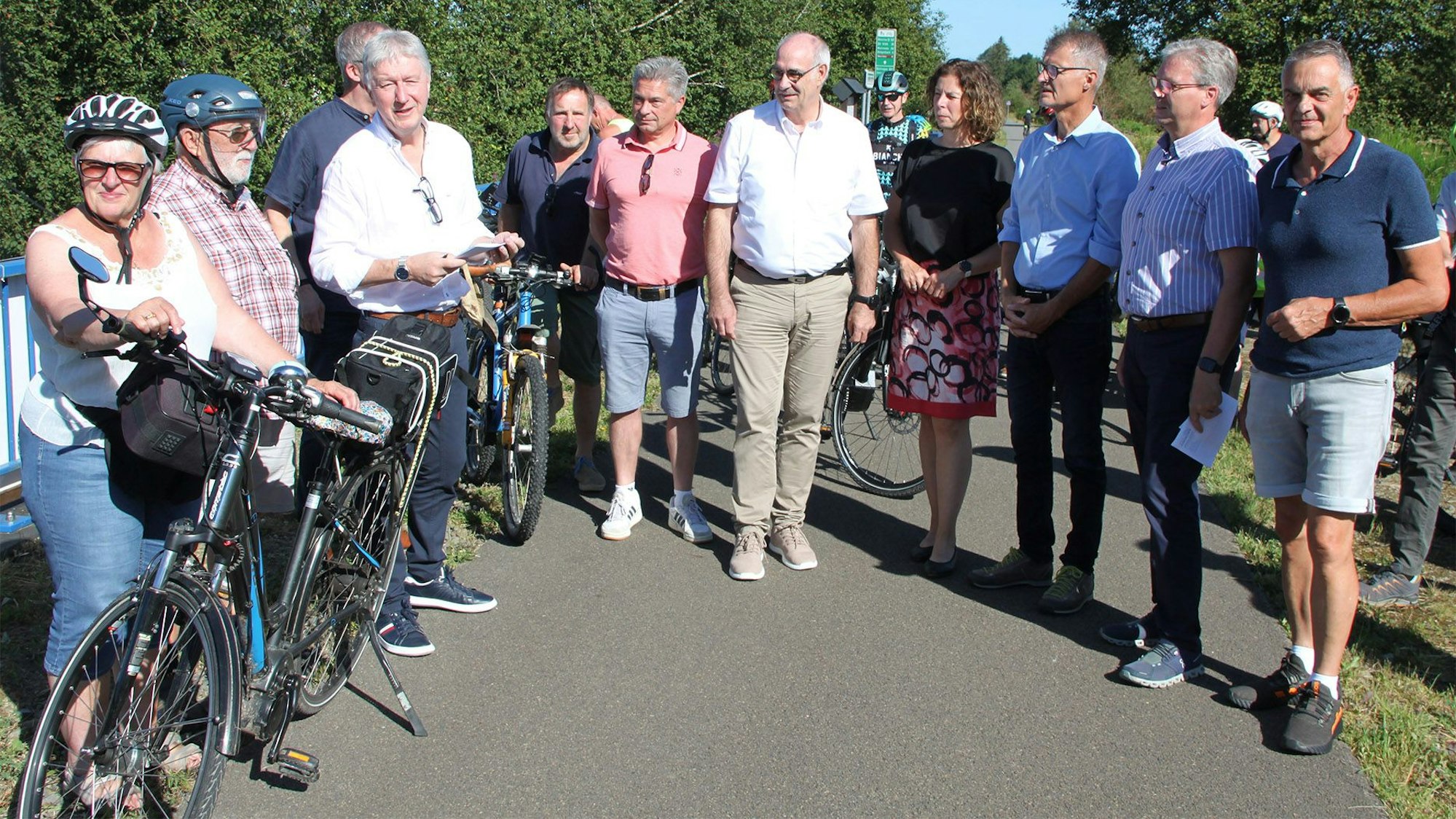 Einige Offizielle und mehrere Radfahrer stehen an einem Sommertag in Freizeitkleidung auf einem Radweg beisammen.